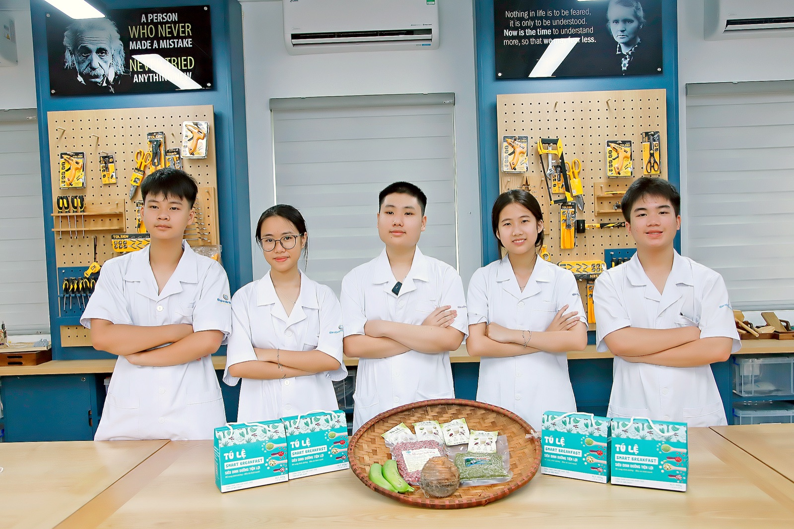 Năm học sinh mặc áo blouse trắng đứng cạnh nhau, phía trước là các hộp Tú Lệ Smart Breakfast và một khay nguyên liệu